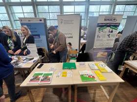 Stół ze stoisku informacyjnym z ulotkami i broszurami, za nim banery organizacji; obok stoją uczestnicy wydarzenia w sali z dużymi oknami.