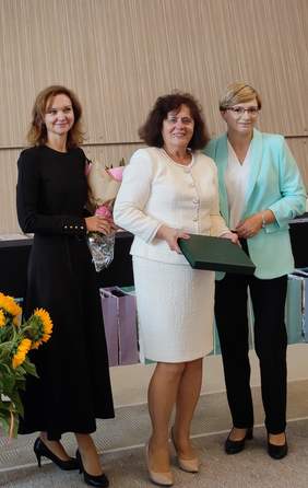 Pokaż zdjęcie: Trzy kobiety stoją razem podczas uroczystości jubileuszowych. Jedna z nich trzyma kwiaty, druga zielone pudełko – Grawerton.