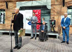 Pokaż zdjęcie: Mężczyzna trzymajacy nagrodę przemawia do mikrofonu. W tle trzech mężczyzn wręczajacych nagrodę, w tym jeden na wózku inwalidzkim.