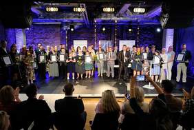 Pokaż zdjęcie: Na scenie stoi duża liczba osób, pozują do pamiątkowego zdjęcia trzymając w ręku dyplomy.