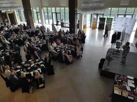 Pokaż zdjęcie: Sala konferencyjna, przy okrągłych stolikach siedzą zaproszeni goście. Wszyscy patrzą w kierunku sceny, na której stoi pani prezes, kobieta w beżowo czarnej sukience..