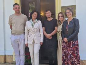 Pokaż zdjęcie: Pięć osób stoi przed drzwiami do budynku - Zakładu Aktywności Zawodowej "U Pana Cogito". Wszyscy pozujący do zjęcia (dwóch mężczyzn i trzy kobiety) są uśmiechnięci. Mężczyźni są pracownikami ZAZ.