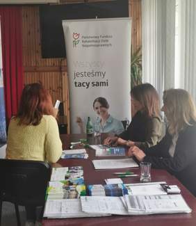 Pokaż zdjęcie: po lewej stronie stołu siedzi tyłem kobieta z żółtym swetrze po prawej stronie dwie kobiety udzielają informacji. Na stole leżą ulotki i informatory, przy stole stoi baner z logo PFRON.