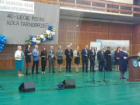 grupa kilkunastu osób stoi  z różami w dłoni, przed nimi kobieta przy mównicy przemawia