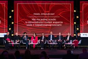 Pokaż zdjęcie: Panel dyskusyjny na scenie konferencyjnej. Kilkoro panelistów siedzi w rzędzie na fotelach, przed nimi stoliki z mikrofonami i wodą. W tle czerwony ekran z napisem: „Panel dyskusyjny. Nie ma jednej ścieżki: o różnorodności modeli wsparcia osób z niepełnosprawnościami”.