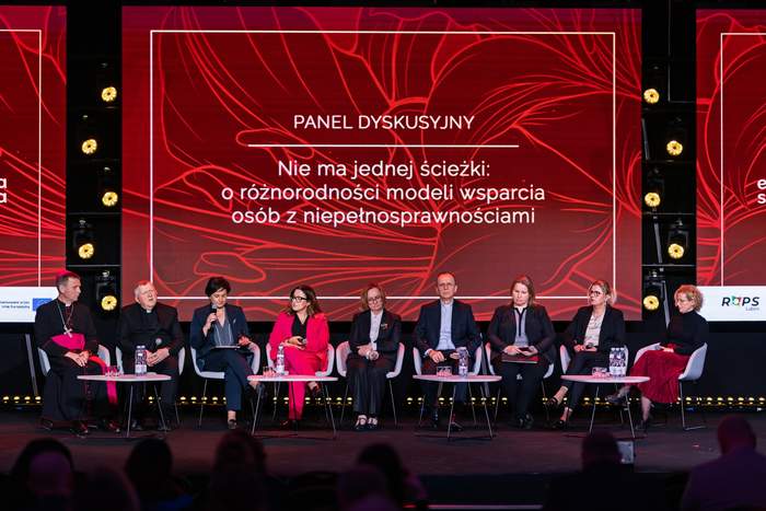 Pokaż zdjęcie: Panel dyskusyjny na scenie konferencyjnej. Kilkoro panelistów siedzi w rzędzie na fotelach, przed nimi stoliki z mikrofonami i wodą. W tle czerwony ekran z napisem: „Panel dyskusyjny. Nie ma jednej ścieżki: o różnorodności modeli wsparcia osób z niepełnosprawnościami”.