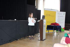 Pokaż zdjęcie: Kobieta z długimi włosami, w białej bluzce i długiej ciemnej spódnicy stoi przy mównicy, prelegent.