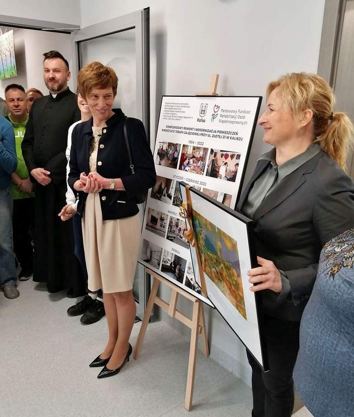 Pokaż zdjęcie: Grupa siedmiu osób, w tym mężczyzn i kobiet stoi na korytarzu oraz w drzwiach prowadzących do jednego z pomieszczeń warsztatu terapii zajęciowej. Kobieta po prawej stronie trzyma w dłoniach obraz. Wśród zgromadzonych osób jest także ksiądz. Przy ścianie ustawiona została sztaluga, na której możemy dostrzec kolaż w antyramie przedstawiający WTZ w Kaliszu, zarówno przed, jak i po kompleksowej modernizacji.