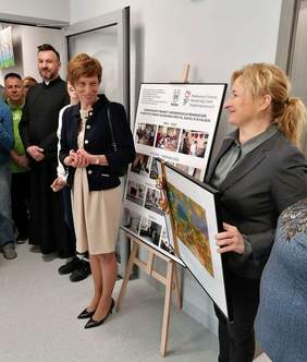Pokaż zdjęcie: Grupa siedmiu osób, w tym mężczyzn i kobiet stoi na korytarzu oraz w drzwiach prowadzących do jednego z pomieszczeń warsztatu terapii zajęciowej. Kobieta po prawej stronie trzyma w dłoniach obraz. Wśród zgromadzonych osób jest także ksiądz. Przy ścianie ustawiona została sztaluga, na której możemy dostrzec kolaż w antyramie przedstawiający WTZ w Kaliszu, zarówno przed, jak i po kompleksowej modernizacji.