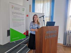 Pokaż zdjęcie: Przy mównicy stoi kobieta w biało niebieskiej bluzce i długiej spódnicy, uśmiecha się do zdjęcia.