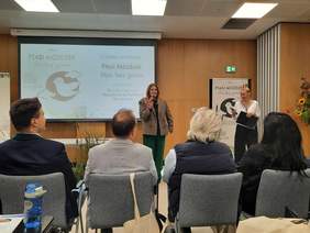 Pokaż zdjęcie: W sali przed widownią stoją dwie kobiety. Trzymają w dłoniach mikrofony i przemawiają do zebranych. Za ich plecami wisi tablica multimedianla z tematem konferencji. 