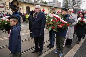 Pokaż zdjęcie: W kolejce stoją ludzie. Trzymają w dłoniach biało-czerwone wiązanki kwiatów. Wśród nich znajduje się dyrektor Łódzkiego Oddziału PFRON. Przed nim stoją harcerze. 