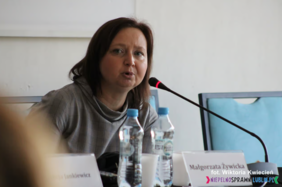 Pokaż zdjęcie: Za biurkiem siedzi kobieta, przed nią stoi mikrofon oraz plakietka z imieniem i nazwiskiem.