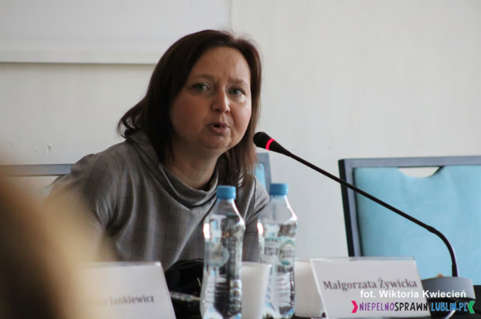 Pokaż zdjęcie: Za biurkiem siedzi kobieta, przed nią stoi mikrofon oraz plakietka z imieniem i nazwiskiem.