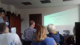 Pokaż zdjęcie: Przedstawiciel Fundacji prezentuje zagadnienia tyflografiki, magnigrafiki oraz audiodeskrypcji. Uczestnicy patrzą na prezentacją wyświetlaną na ścianie.