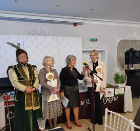 Pokaż zdjęcie: Cztery osoby stoją na scenie podczas uroczystości wręczenia nagród. Dwie kobiety trzymają w rękach dekoracyjne statuetki. Kobieta z mikrofonem przemawia, a po lewej stronie stoi mężczyzna w bogato zdobionym stroju szlacheckim z piórami w czapce.