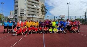 Pokaż zdjęcie: Grupowe zdjęcie uczestników turnieju piłkarskiego „Futsal Bez Barier” – zawodnicy w kolorowych koszulkach oraz sędziowie pozują na boisku sportowym przy szkole w Białymstoku. W tle widać budynki mieszkalne i kosze do koszykówki.