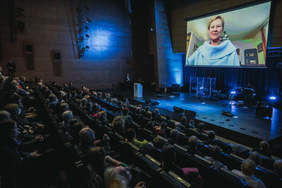 Pokaż zdjęcie: Duża sala teatralna, w której siedzi wiele osób ubranych na galowo, nad scena oświetloną na niebiesko wisi potężny telebim na którym wyświetlany jest film z przemawiającą kobietą.