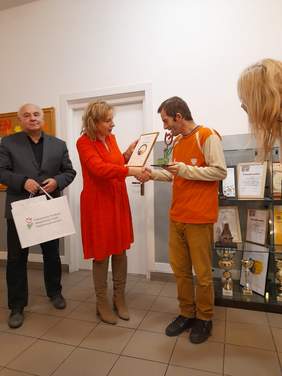 Pokaż zdjęcie: Kobieta, blondynka w czerwonej sukience wręcza dyplom i składa gratulacje uczestnikowi konkursu