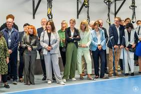 Pokaż zdjęcie: W hali sportowej znajduje się grupa kilkunastu osób, która ustawiona jest w dwóch rzędach. Większość osób ma skierowany wzrok w jedną stronę.