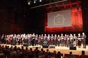 Pokaż zdjęcie: Na scenie Filharmonii Łódzkiej stoją w rzędzie ludzie. Wszyscy trzymają w dłoniach kwiaty i odznaczenia. Widać również osoby siedzące na widowni. 