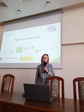 Pokaż zdjęcie: Prelekcję prowadzi kobieta w szarej sukience z jasnymi półdługimi włosami.