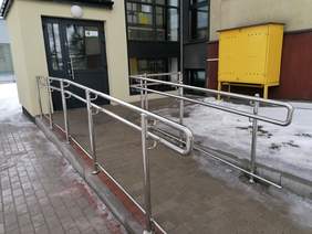 Pokaż zdjęcie: podjazd prowadzący do drzwi wejściowych wykonany z kostki betonowej. Po obu stronach stalowe balustrady z poręczami