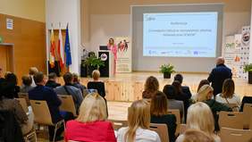 Pokaż zdjęcie: Kobieta stoi na podeście przemawia do osób siedzących naprzeciwko za nią na ekranie wyświetla się napis Konferencja O oswajaniu inkluzji w rzeczywistości szkolnej – doświadczenia SCWEW.