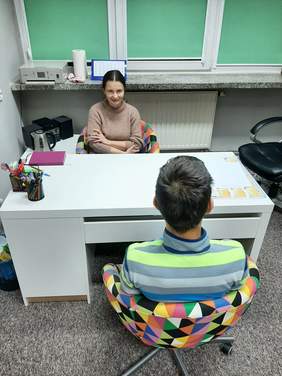 Pokaż zdjęcie: Przy biurku na fotelu siedzi uśmiechnięta młoda kobieta, po przeciwnej stronie biurka siedzi młody chłopak.
