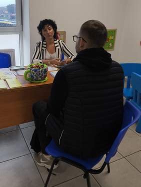 Pokaż zdjęcie: Przy biurku rozmawiają kobieta z mężczyzną, na stole leżą ulotki.