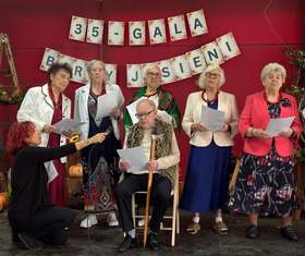 Grupa starszych osób występuje na scenie podczas „35 - Gali Barwy Jesieni”. Na środku siedzi starszy, jasnowłosy mężczyzna w okularach, jasnej koszuli, wzorzystej brązowo-złotej kamizelce i czarnych spodniach, trzymający w dłoniach kartkę. Przed nim klęczy osoba w czarnym stroju, kierując w jego stronę mikrofon. Po obu stronach mężczyzny stoją elegancko ubrane starsze kobiety w żakietach, spódnicach i sukienkach, które trzymają kartki i śpiewają.