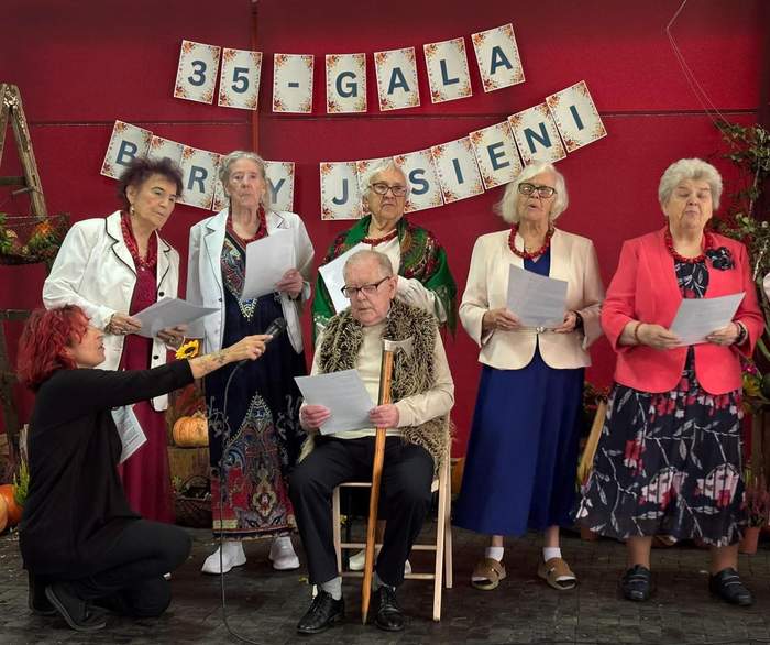 Pokaż zdjęcie: Grupa starszych osób występuje na scenie podczas „35 - Gali Barwy Jesieni”. Na środku siedzi starszy, jasnowłosy mężczyzna w okularach, jasnej koszuli, wzorzystej brązowo-złotej kamizelce i czarnych spodniach, trzymający w dłoniach kartkę. Przed nim klęczy osoba w czarnym stroju, kierując w jego stronę mikrofon. Po obu stronach mężczyzny stoją elegancko ubrane starsze kobiety w żakietach, spódnicach i sukienkach, które trzymają kartki i śpiewają.
