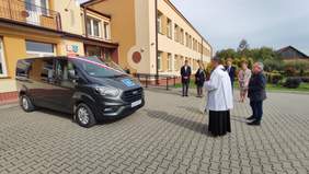 Pokaż zdjęcie: Kobiety i mężczyźni elegancko ubrani patrzą w kierunu szarego samochodu - busa przystosowanego do przewozu osób z niepełnosprawnościami