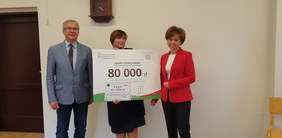 Pokaż zdjęcie: trzy osoby stoją obok siebie. Dwie kobiety trzymają dużą tablicę symbolizującą czek. 