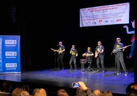 Pokaż zdjęcie: Na scenie pięciu mężczyzn ubranych na czarno. Jeden z nich śpiewa, drugi gra na perkusji, trzech gra na gitarze. Nad nimi baner z napisem  XXVII Przegląd Twórczości Artystycznej Osób Niepełnosprawnych Białystok 2024.