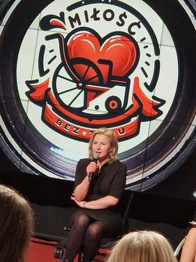 Pokaż zdjęcie: Kobieta, blondynka, ubrana w czarną krótką sukienkę, siedzi na krześle stojącym przed publicznością. W ręku trzyma mikrofon. 