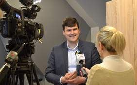 Pokaż zdjęcie: Mężczyzna stoi przed kamerą i udziela wywiadu. Obok niego stoi kobieta, która trzyma mikrofon