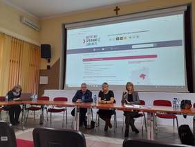 Pokaż zdjęcie: Sala konferencyjna w tle duży ekran, przed ekranem duży stół prezydialny, przy którym siedzą cztery kobiety, zdjęcie zrobione od strony widowni