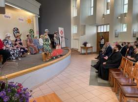 Pokaż zdjęcie: Po lewej stronie scena, na której stoi starsza kobieta w kapeluszu i kwiecistej sukience, za nią siedzi na krzesłach kilka kobiet podobnie ubranych, po prawej stronie widownia, w pierwszym rzędzie siedzą księża. W tle logo Caritas.