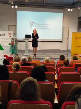 Pokaż zdjęcie: Zdjęcie z tyłu sali w kierunku sceny. Na scenie kobieta blondynka w czarnej sukience.