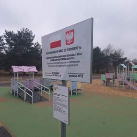 Pokaż zdjęcie: Dwa wielofunkcyjne urządzenie zabawowe, jedno na nawierzchni z piasku, a drugie na lekko utwardzonej. Z przodu ustawiona jest tablica informująca o dofinansowaniu inwestycji ze środków PFRON. W tle znajdują się drzewa różnej wielkości.