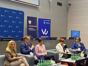 Pokaż zdjęcie: Prelegenci uczestniczący w panelu dyskusyjnym siedzą na czterech sofach ustawionych w rzędzie. Wśród panelistów są trzy kobiety oraz dwóch mężczyzn. Jeden z nich, w okularach i garniturze, przemawia do zgromadzonych osób, trzymając w ręce mikrofon.