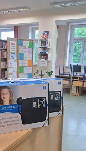 Pokaż zdjęcie: Mobilne pętle indukcyjne zapakowane w kartonie ustawione są na biurku. W tle pomieszczenia z oświetleniem LED znajduje się regał z książkami, tablica korkowa z przyczepionymi do niej kartkami oraz biurko z dwoma położonymi na nim krzesłami.