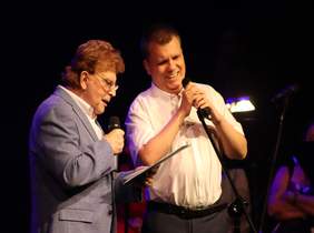 Pokaż zdjęcie: w tle ciemna scena na której stoją dwaj mężczyźni i spiewają do mikrofonu 