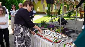 Pokaż zdjęcie: Elegancko ubrana kobieta kroi tort. Na torcie jest napis: Dom Pomocy Społecznej w Wiśniowej Górze. W tle widać inne osoby. 
