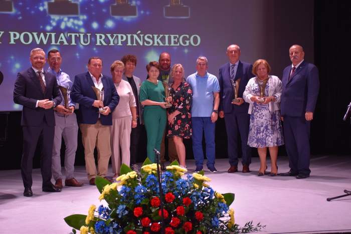 Pokaż zdjęcie: Grupa osób stoi na scenie, kiloro trzyma otrzymane statuetki. Na pierwszym planie stoi kosz z czerwono-niebiesko-żółtymi kwiatami.