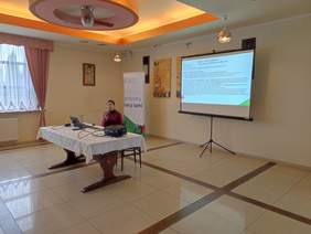 Pokaż zdjęcie: Przy stole ma którym jest laptop siedzi kobieta w w dłoniach trzyma mikrofon. Za nią na ekranie wyświetlona jest prezentacja.