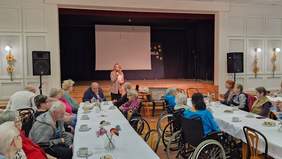 Pokaż zdjęcie: Kobieta z mikrofonem przemawia do mieszkańców Domu Pomocy Społecznej w Lisówkach. Po bokach sceny z białym ekranem stoją głośniki, a przy długich stołach siedzą słuchający uczestnicy.