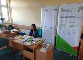 Stoisko informacyjne PFRON, pzry biurku siedzi pracownik PFRON, na którym leżą materiały promocyjne, z tyłu widać banery z logo PFRON i CIDON
