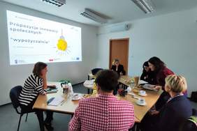 Pokaż zdjęcie: Kilka kobiet siedzi przy stolikach i rozmawiają. Na ścianie sali wyświetlana jest prezentacja z innowacjami.